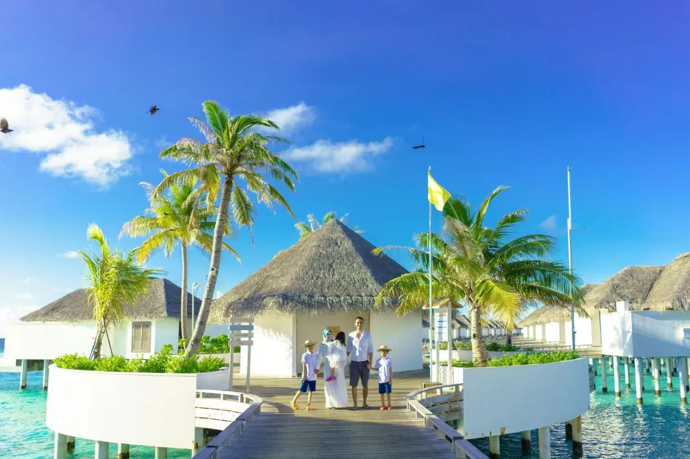 Family of four on a boardwalk between thatched overwater villas, turquoise water below and a bright blue sky.