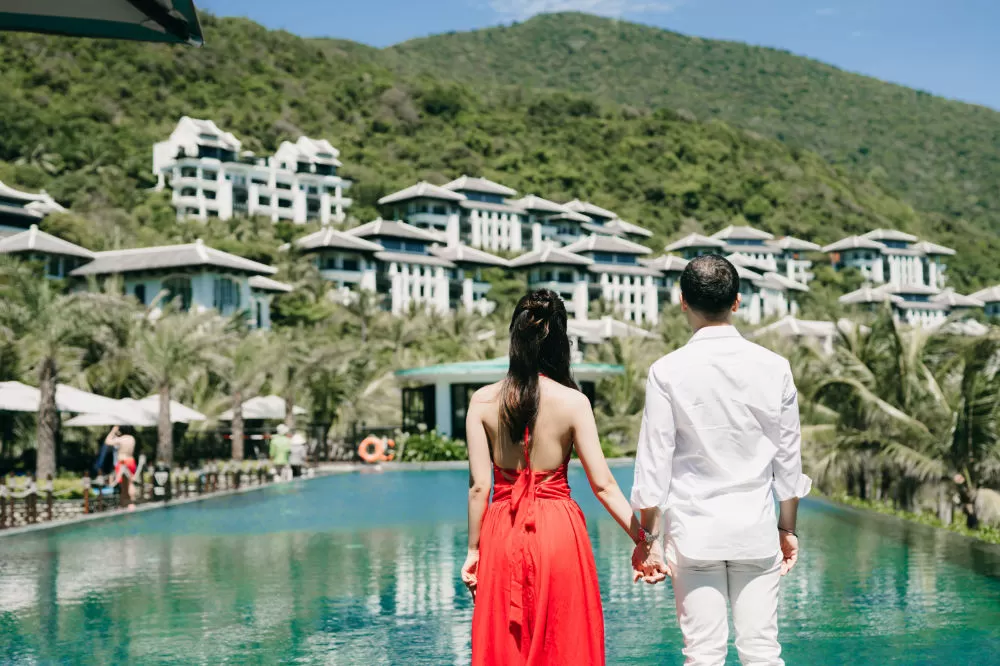 Couple holding hands at the edge of a pool, looking toward a white hillside resort set among green mountains.