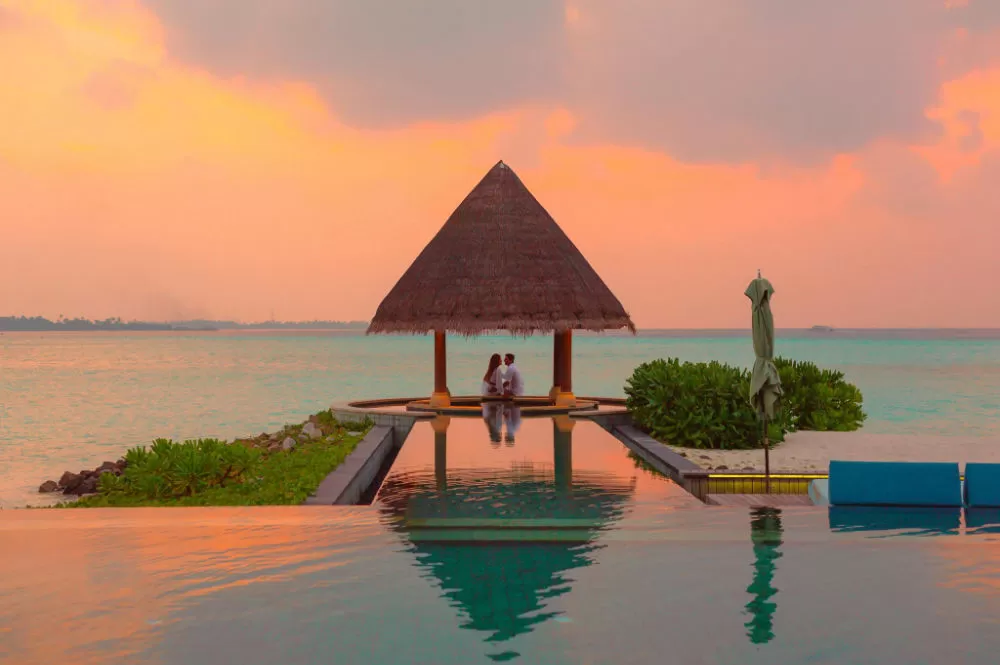 Couple seated beneath a thatched gazebo at the edge of an infinity pool, facing the sea under an orange sunset.