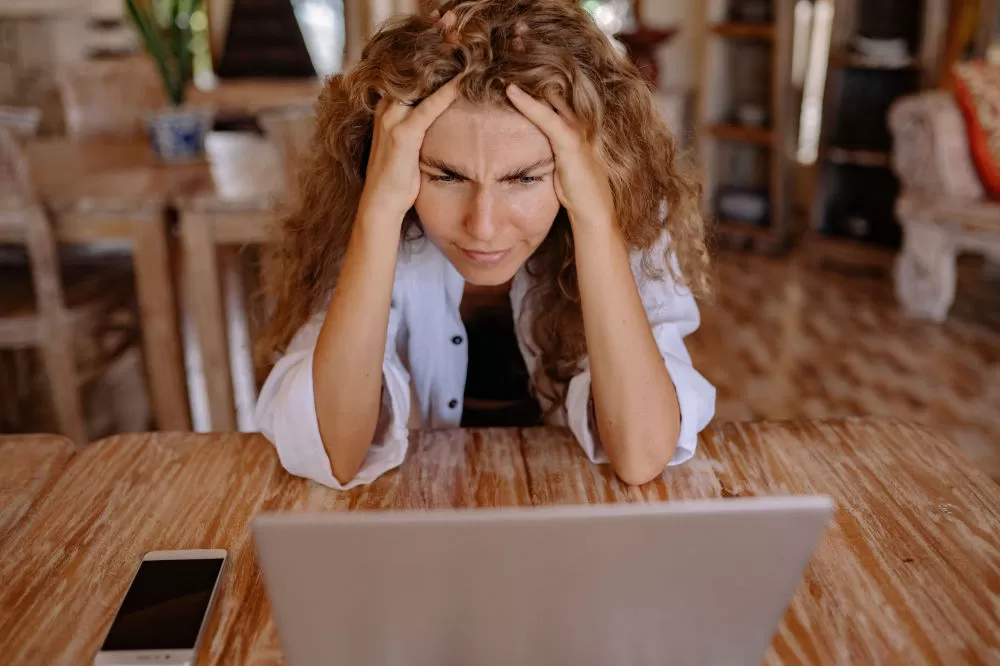 Frustrated woman with her head in her hands, frowning at a laptop on a wooden table.