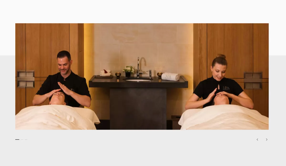Spa gallery slide: two therapists give facials to guests lying side by side in a warm, wood-panelled suite.