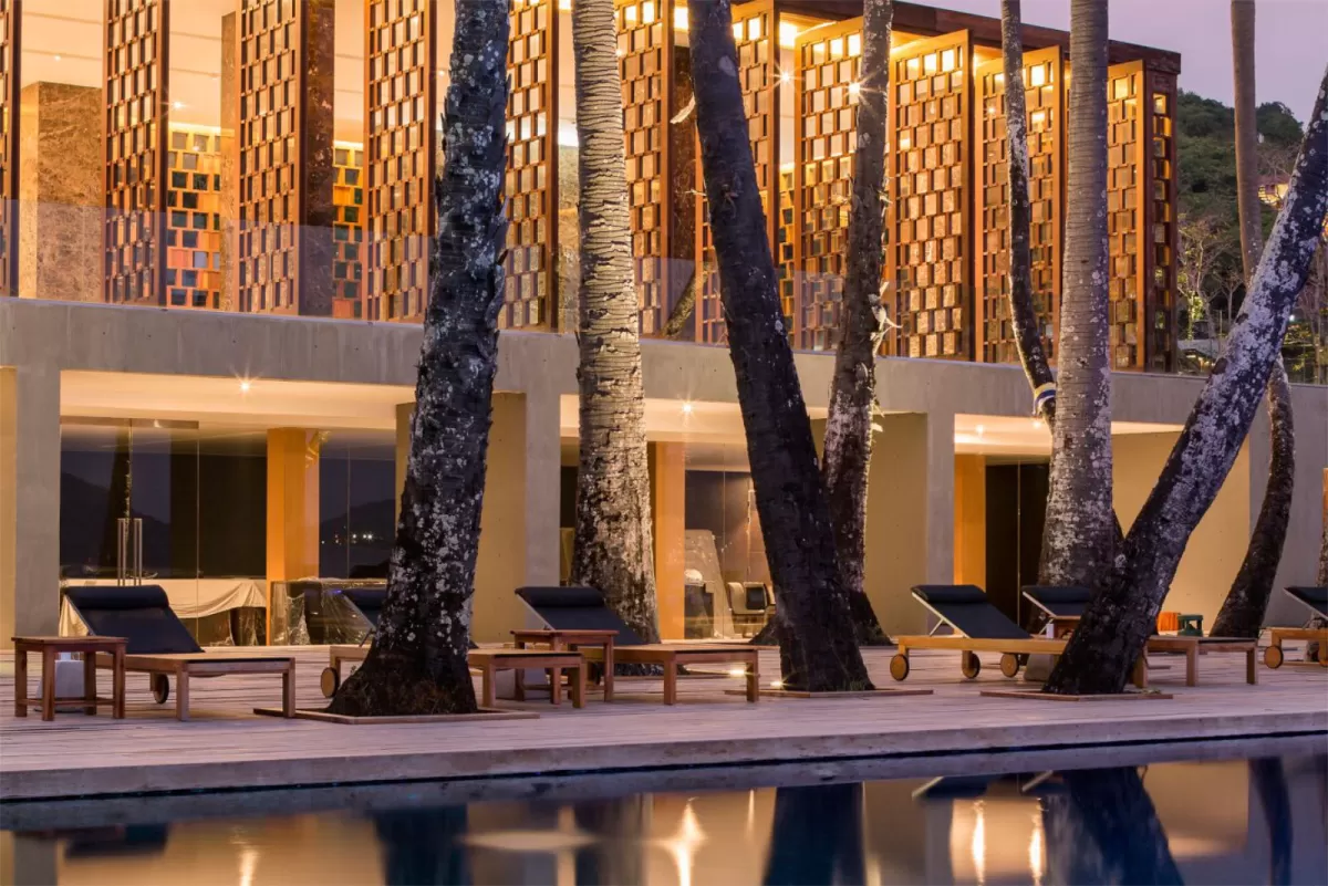 Resort pool deck at dusk, loungers set between palm trunks before a glowing latticed facade.