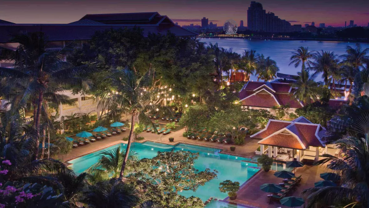 Riverside resort at dusk: a lit pool and Thai-style pavilions among palms, with city towers across the river.