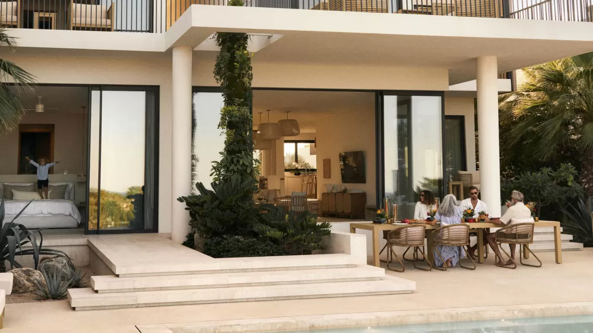 A multigenerational family enjoys an outdoor dinner on a sunny patio at an Altaris resort villa next to a swimming pool, while a young boy plays on a bed inside an open-concept suite in the background.
