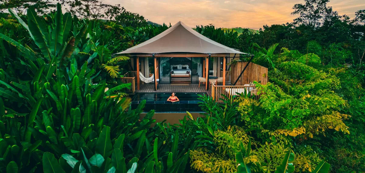 An expansive aerial view of an eco-luxury tented resort village nestled in a lush green valley forest, with a prominent volcanic mountain partially shrouded in clouds rising in the background.