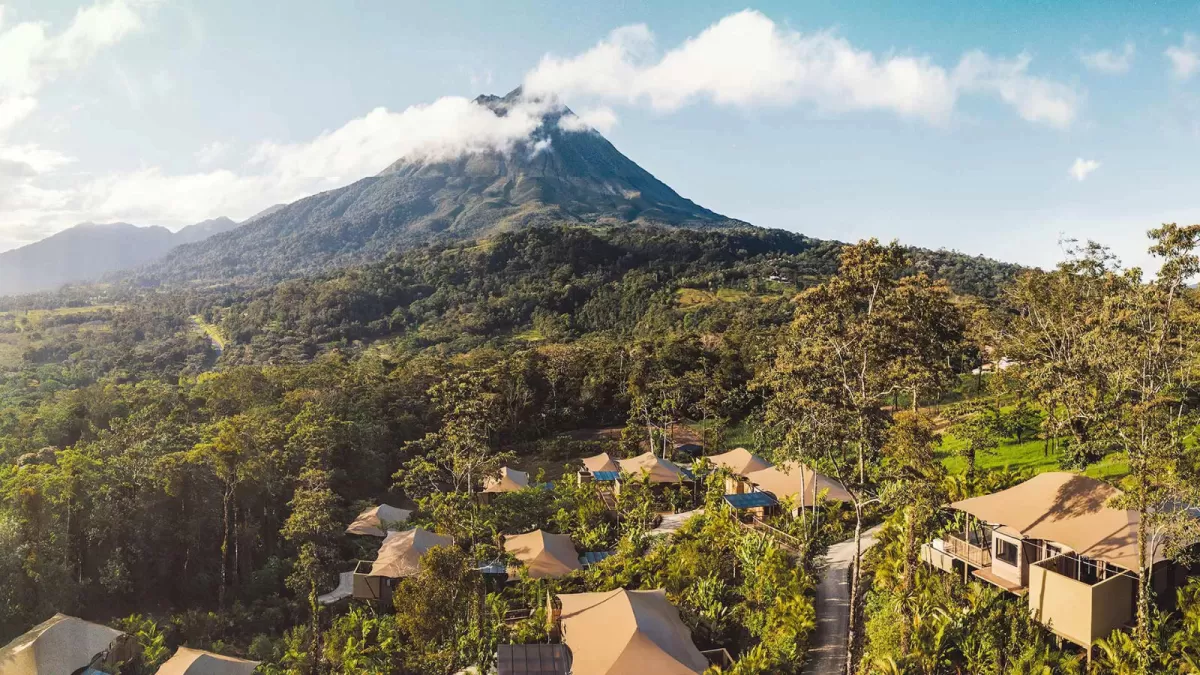 High-angle view of a luxury safari tented villa hidden in a dense tropical jungle at sunset, featuring a private plunge pool where a woman rests at the edge, an open deck with a hammock, and a visible bedroom suite inside.