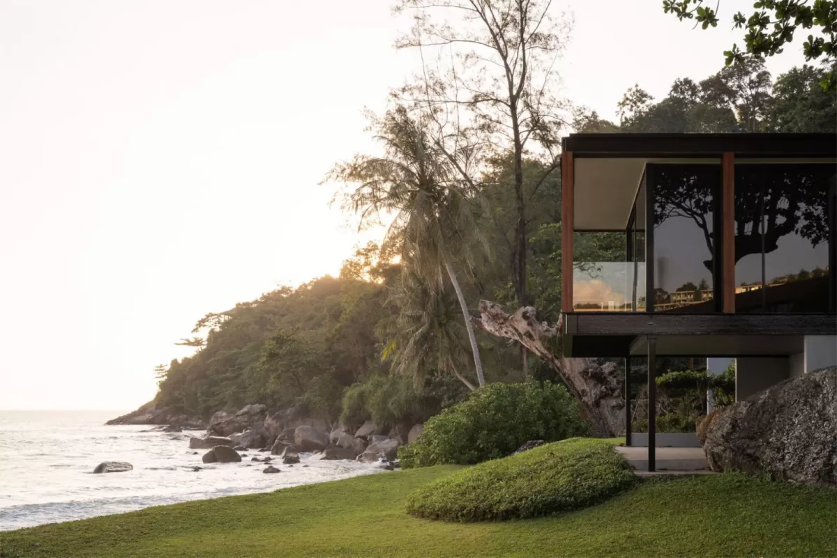 An elevated, modern minimalist glass-walled resort pavilion set on a rocky shoreline cliff surrounded by tropical palm trees and greenery at sunset, overlooking the ocean.