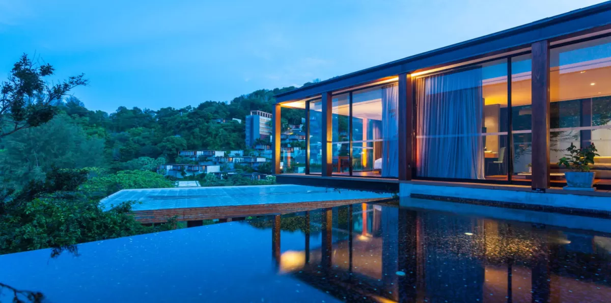 Infinity pool reflecting a modern luxury villa with floor-to-ceiling glass windows at dusk, Della Resort.