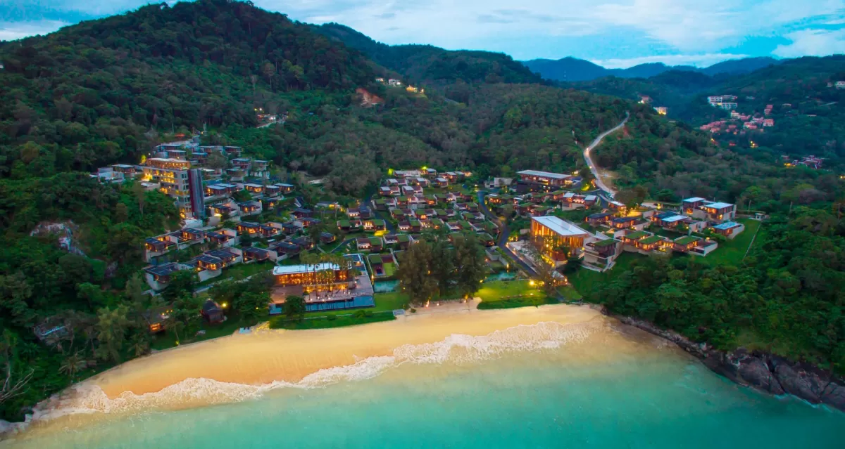 Aerial twilight view of a coastal luxury resort nestled in a lush hillside forest bordering a white sand beach and turquoise water.